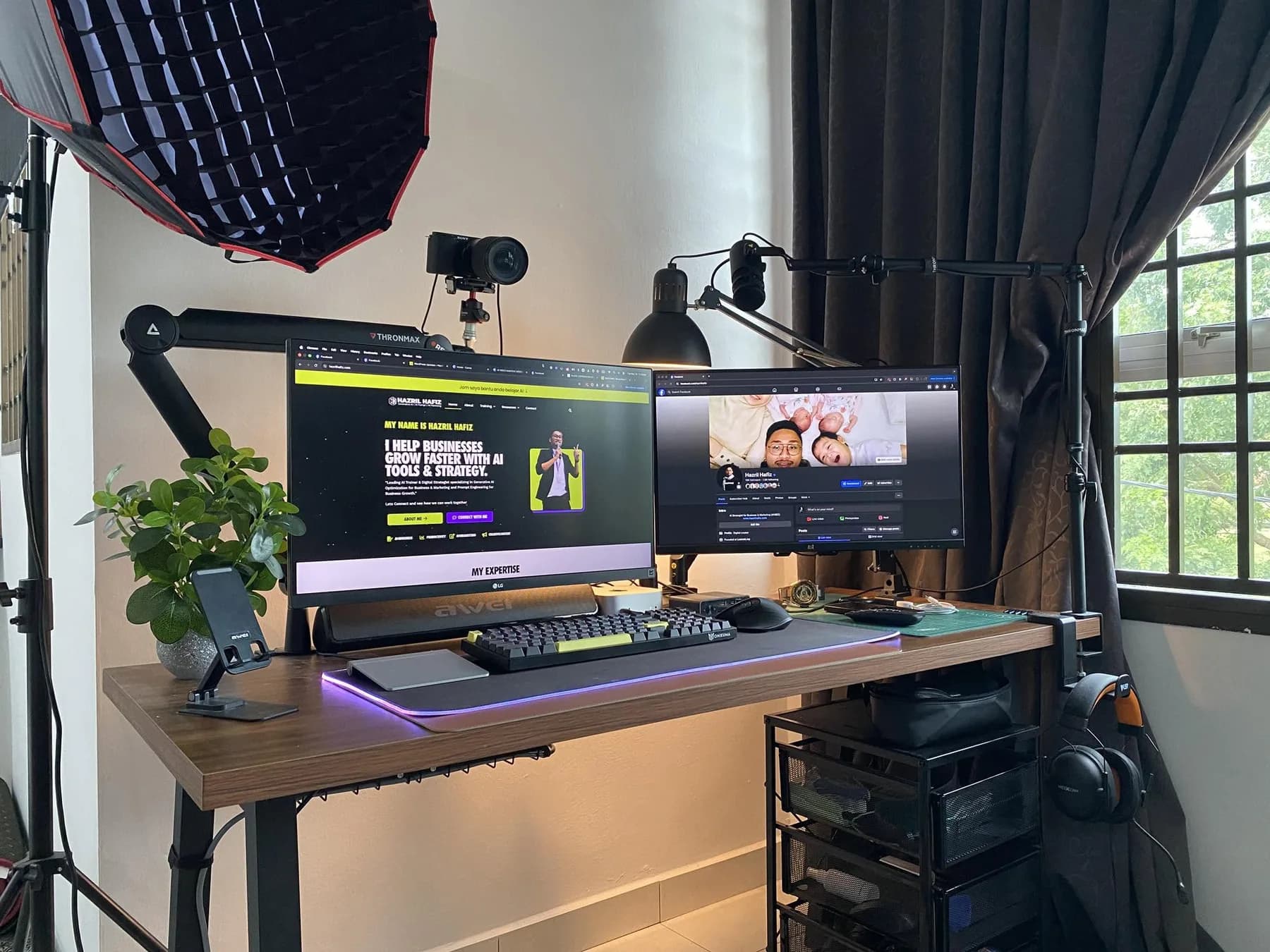 A wider view of the same desk: the LG monitor with hazrilhafiz.com open, an ultrawide monitor next to it, an RGB-lit desk mat, a mechanical keyboard, and my headphones resting on the trolley, with the edge of the softbox just in frame on the left.