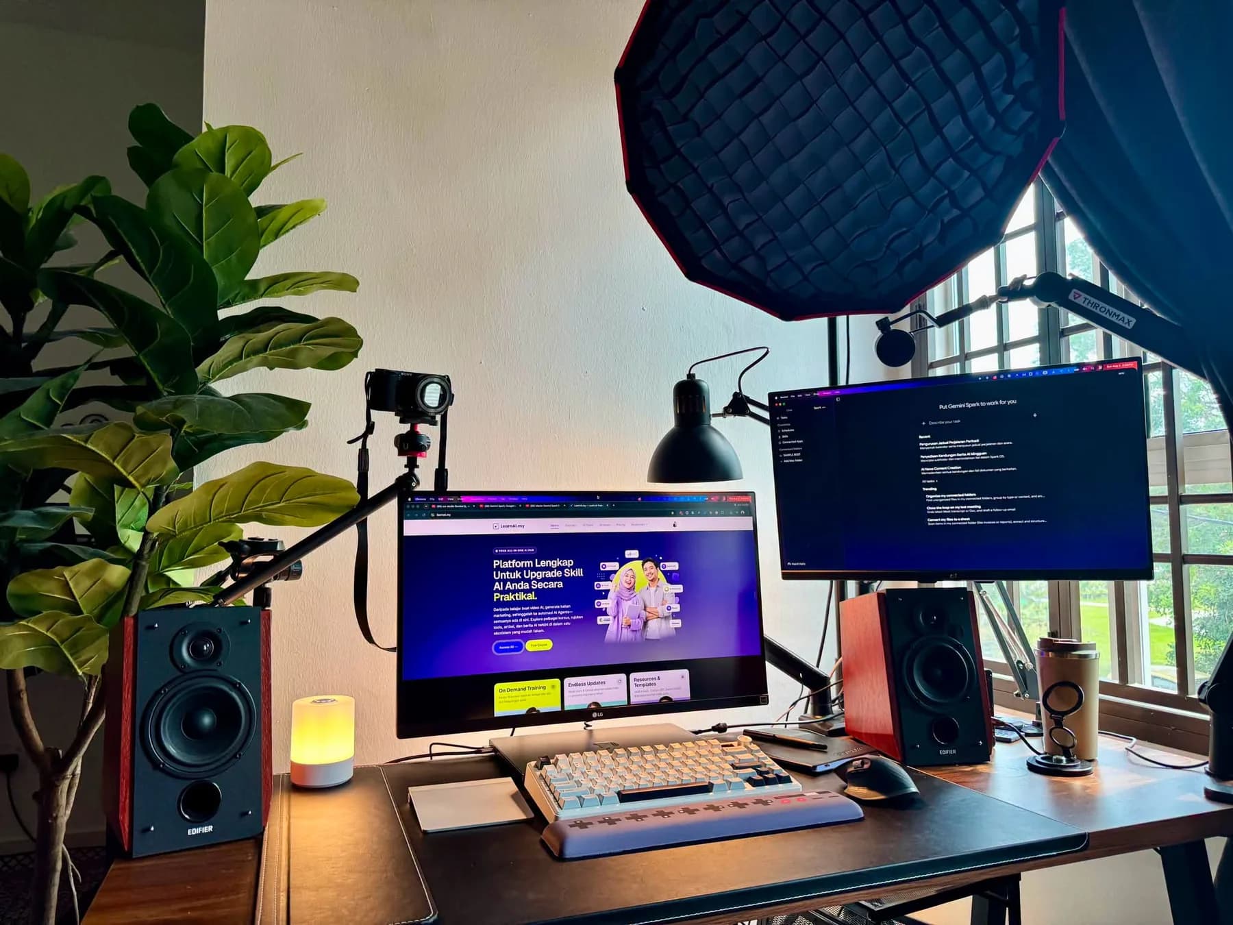 My desk photographed head-on: an LG monitor with LearnAI.my open on it, a second monitor on an arm beside it, Edifier bookshelf speakers standing either side of the desk, a softbox lighting the room from above, my Sony ZV-E10 on a desk mount and a Thronmax boom arm swung in from the right, with a fiddle-leaf plant in the left corner.